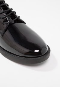 Chaussure en cuir verni noir avec un bout arrondi, un design à lacets et une semelle en caoutchouc. Présente une surface lisse et réfléchissante ainsi que des accents brodés.
