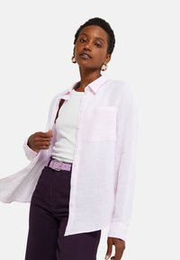 Chemise boutonnée en lin rose clair avec une poche poitrine, portée sur un débardeur blanc et un pantalon taille haute violet foncé, accompagné d'une ceinture assortie.