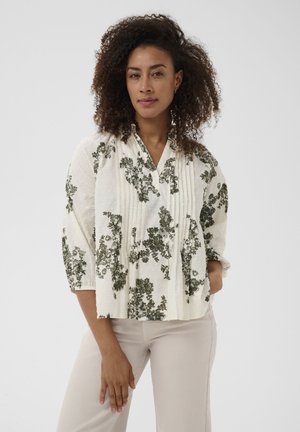 Žena s kučeravými vlasmi nosí bielu blúzku so zeleným kvetinovým vzorom a svetlo béžové nohavice, stojaca pred jednoduchým pozadím.