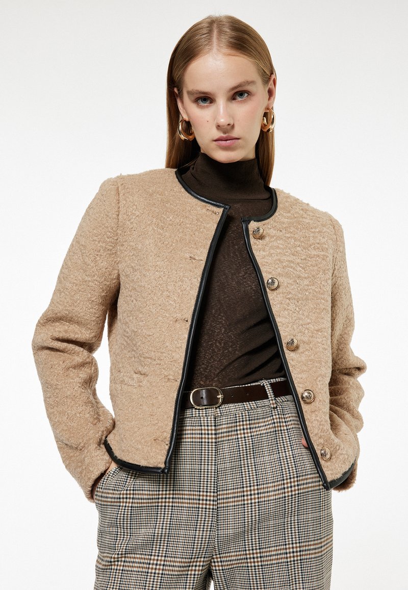 Beżowa kurtka o fakturze z czarnymi wykończeniami, okrągłym dekoltem i guzikami z przodu, połączona z brązowym golfem i w kratkę spodniami.