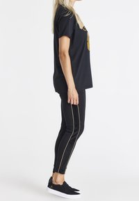 Černé tričko s krátkým rukávem a zlatou grafikou, spárované s černými legínami se zlatými bočními pruhy a černými slip-on teniskami.
