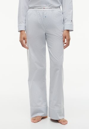 Persona che indossa pantaloni del pigiama a righe bianchi e azzurri chiari e maglia a maniche lunghe coordinata, in piedi a piedi nudi su sfondo bianco.
