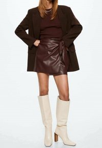 Gonna avvolgente in pelle marrone, top ribbed color maroon, blazer oversize nero e stivali al ginocchio avorio con tacco quadrato.
