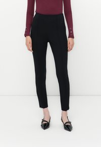 Donna che indossa pantaloni neri aderenti con fessure alla caviglia, abbinati a un top aderente a maniche lunghe bordeaux e scarpe nere eleganti a punta.