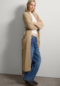 Beige geribbelde gebreide lange cardigan met zakken, gedragen over een witte top en losse blauwe spijkerbroek, voor een neutrale muur.