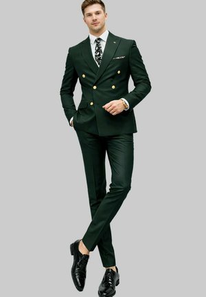 Homme debout, jambes croisées, portant un costume croisé vert foncé, une chemise blanche, une cravate à fleurs, des chaussures habillées noires et une montre-bracelet.