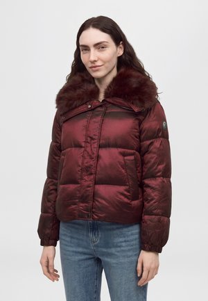 Une femme porte une veste matelassée bordeaux à grand col en fourrure et un jean bleu, sur un fond clair uni.