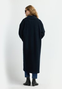 Langer, dunkelblauer Mantel mit vertikalen Nadelstreifen, breitem Kragen und oversized Passform. Getragen über blauen Jeans und schwarzen Stiefeletten.