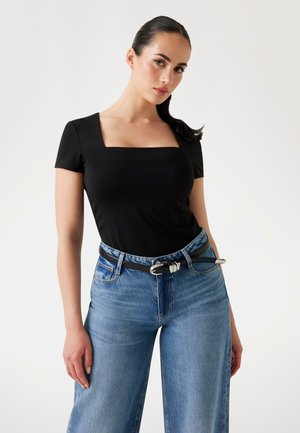 Woman with dark hair in a ponytail wears a black square-neck top, light blue high-waisted jeans, and a black belt with silver buckle.