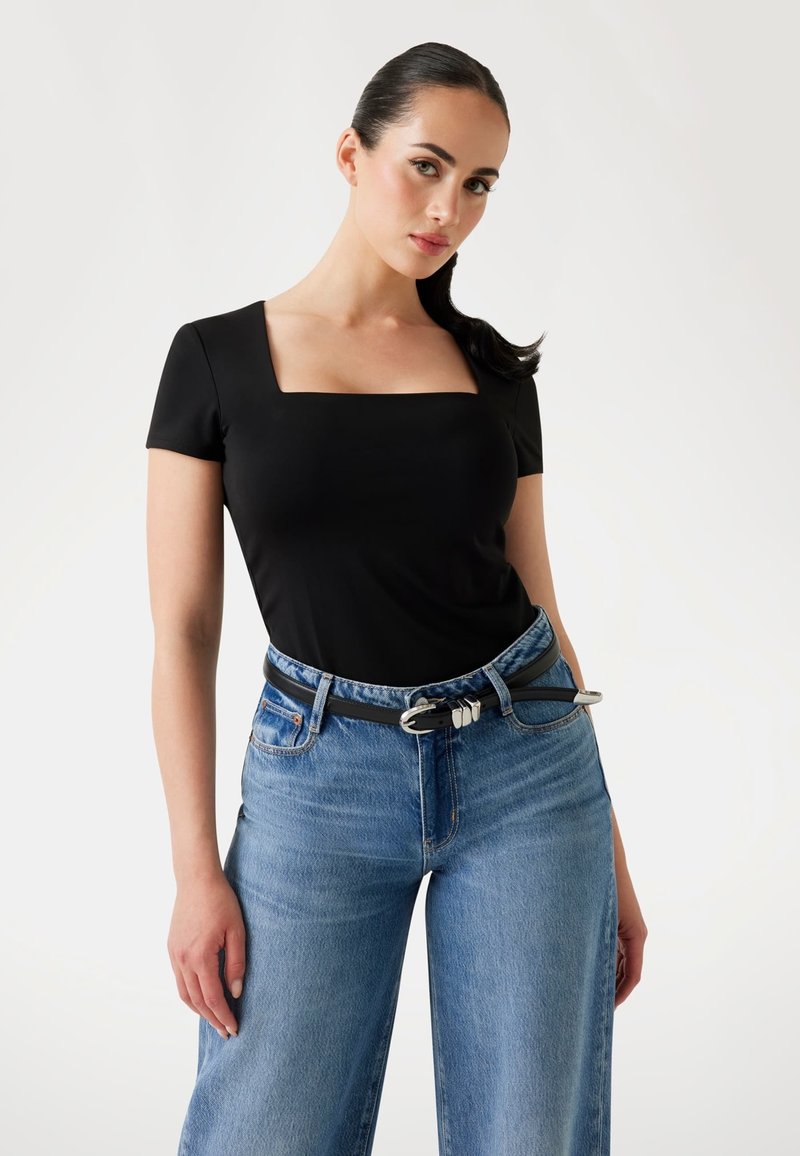 Femme aux cheveux bruns en queue de cheval portant un top noir à encolure carrée, un jean taille haute bleu clair et une ceinture noire avec boucle en argent.