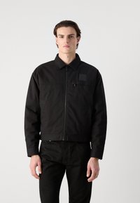 Jachetă neagră scurtă cu guler, fermoar frontal și o eticheta pătrată cu logo pe pieptul stâng. Pur tată peste pantaloni negri. Textură netedă.