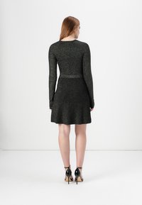 Una modella con capelli rossi lisci indossa un vestito attillato nero scintillante e tacchi alti, mostrata di spalle su uno sfondo bianco semplice.