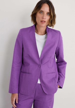 Fialový blazer s vykrojeným límcem, zapínáním na jeden knoflík a dvěma předními kapsami. Nosený přes bílou košili, která má hladkou texturu.