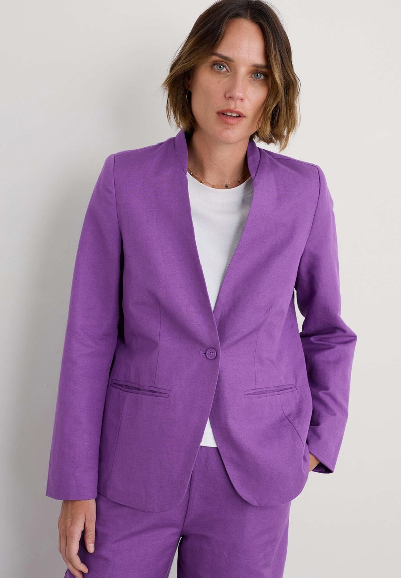 Violetinis blazeris su išpjautu apykaklė, vienu mygtuko uždarymu ir dviem priekinėmis kišenėmis. Dėvimas ant balto marškinio, turinčio švelnų tekstūrą.