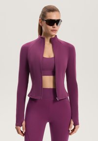 Fialová sportovní bunda s přiléhavým střihem, dlouhými rukávy a předním zapínáním na zip. Kombinována s odpovídajícím crop topem a legínami.