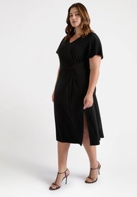 Zwart midi-jurk met een V-hals, korte mouwen en een zijsplit. Gemaakt van gladde stof met een geplooide taille; gecombineerd met hakken.