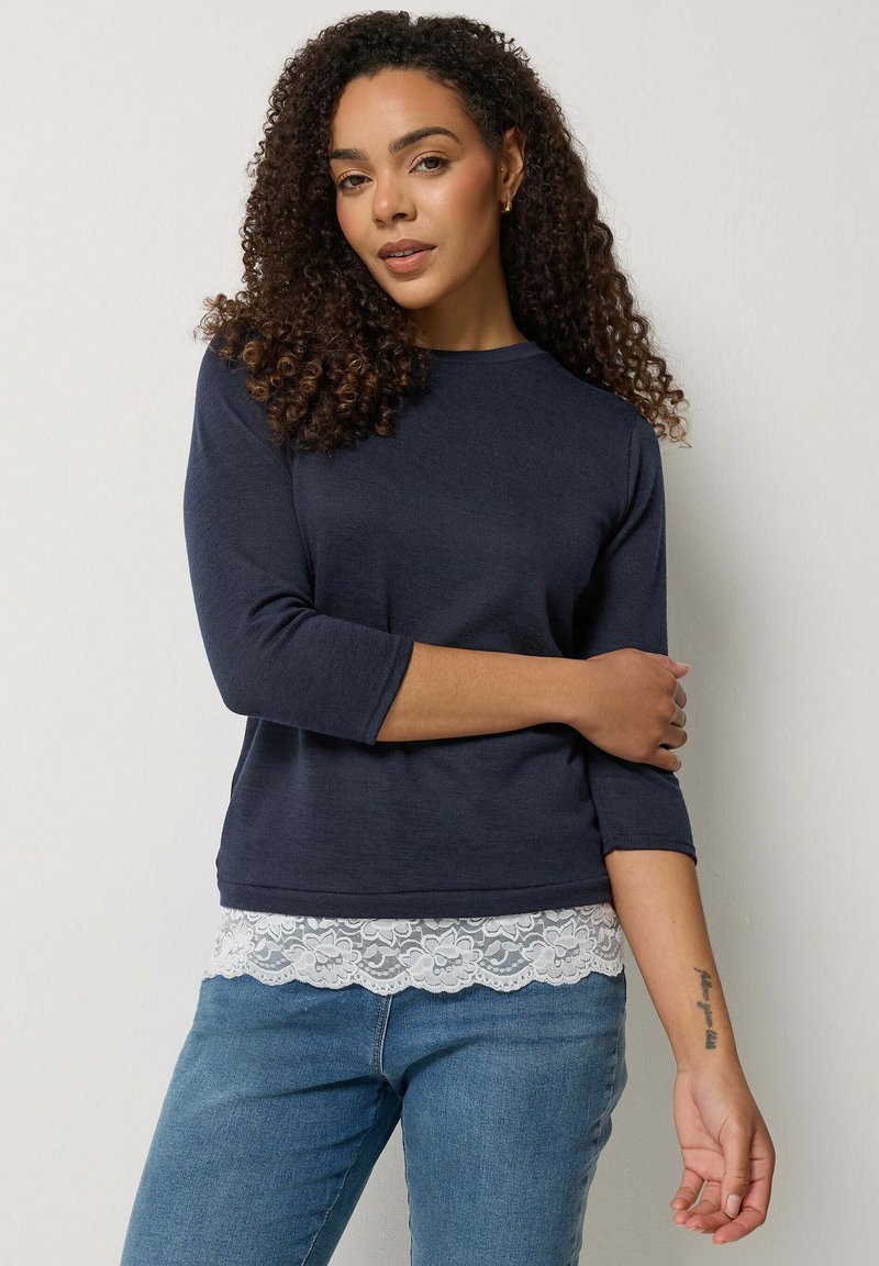 Frau mit lockigem Haar, die ein marineblaues Langarmshirt mit Spitzenbesatz am Saum und blaue Jeans trägt, steht vor einem schlichten hellen Hintergrund.