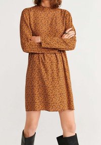 Braun Kleid mit langen Ärmeln, rundem Halsausschnitt und schwarzem Sprenkelmuster. Das Material wirkt weich; A-Linie Silhouette mit einem Taillengürtel.