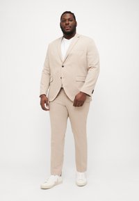 Beige suit with a tailored fit; single-button jacket, two front pockets, and matching tapered pants. Worn with a white t-shirt and light sneakers.