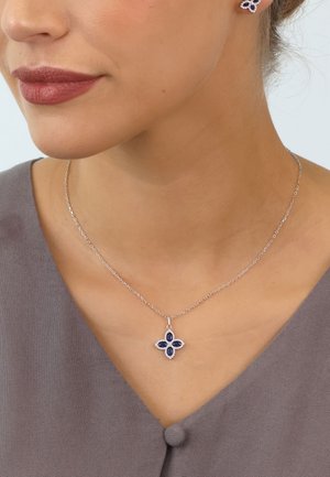 Silberne Kettenhalskette mit einem blumenförmigen Anhänger, der mit blauen Edelsteinen besetzt ist und einem Halo aus kleineren, klaren Steinen. Glatte Metalloberfläche.