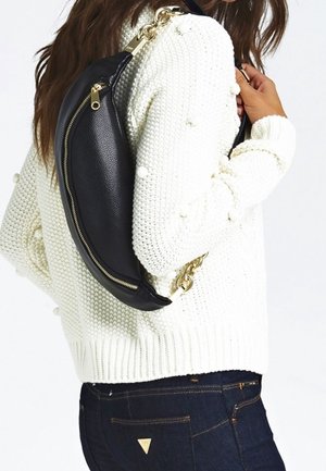 Sac bandoulière en cuir noir avec une forme courbée et des ferrures dorées, porté sur un pull en tricot blanc et assorti à un jean foncé.