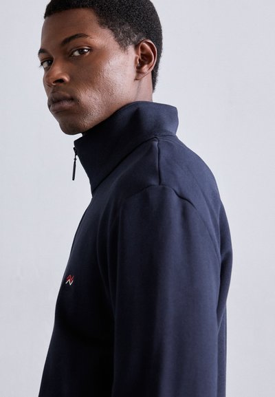 Sweatshirt azul-marinho com fecho de correr e gola alta, apresentando um design minimalista e um pequeno logótipo vermelho e branco no peito. Textura suave.