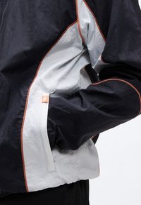 Μανίκι μαύρου και άσπρου μπουφάν με πορτοκαλί ραφές και μια μικρή πορτοκαλί ετικέτα κοντά σε μια τσέπη όπου βρίσκεται ένα λυγισμένο χέρι.