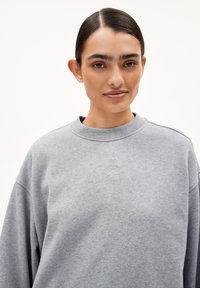 Junge Frau mit nach hinten gekämmtem dunklem Haar, die einen hellgrauen Sweatshirt mit einem kleinen gestickten Design auf der Brust trägt, vor neutralem Hintergrund.