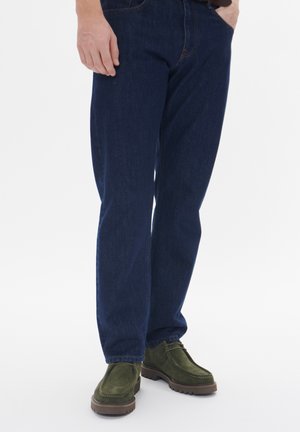 Blugi de culoare bleumarin, cu croială dreaptă, asortați cu pantofi din piele de căprioară verzi, cu talpă din cauciuc. Design simplu, cu detalii de cusătură vizibile.