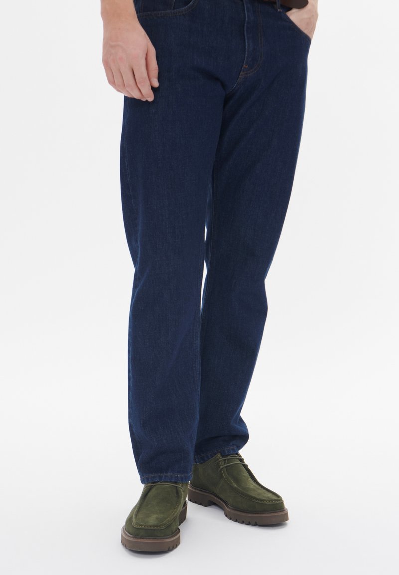 Blugi de culoare bleumarin, cu croială dreaptă, asortați cu pantofi din piele de căprioară verzi, cu talpă din cauciuc. Design simplu, cu detalii de cusătură vizibile.