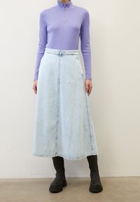 Jupe maxi en denim bleu clair à taille haute, associée à un haut ajusté à manches longues lavande avec un col zippé. Bottes noires épaisses.