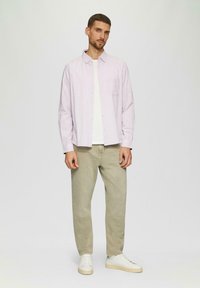 Lange mouw lichtroze overhemden met knopen over een wit T-shirt, gecombineerd met beige broeken en witte sneakers op een neutrale achtergrond.