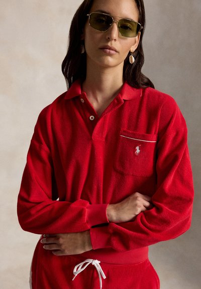 Femme portant des lunettes de soleil à verres verts, des boucles d'oreilles en forme de coquillage, un polo à manches longues rouge avec un logo sur la poche et un pantalon rouge assorti avec un cordon blanc.