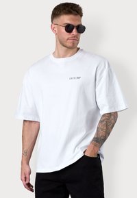 Weißes oversized T-Shirt aus Baumwolle mit kurzen Ärmeln und einem kleinen schwarzen Logo. Das Model trägt dunkle Hosen und runde Sonnenbrillen.