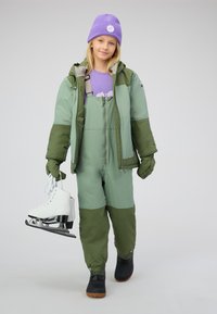 Bambino in abbigliamento invernale verde e cappello viola che tiene in mano pattini da ghiaccio bianchi, in piedi su uno sfondo chiaro uniforme.