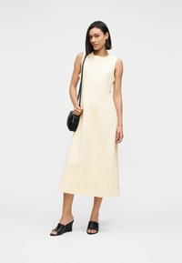 Γυναίκα με αμάνικο κρεμ μίντι φόρεμα με πλευρικά κουμπιά, μαύρα γόνατα με σφήνα και μαύρη τσάντα χιαστί σε απλό φόντο.