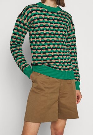 Kobieta ubrana w sweter z długim rękawem w zielono-czarno-beżowy wzór z prążkowanymi mankietami i dołem, do tego brązowe szorty do kolan.