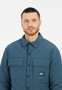 Gequilted jack in teal, met een knoopsluiting aan de voorkant, twee borstzakken en een logo patch. De gestructureerde ruitpatroon versterkt het ontwerp.