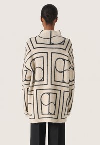 Beige oversized tröja med svart geometriskt mönster och öppen krage. Mjuk textur, långa ärmar och avslappnad passform, kombinerad med svarta byxor.