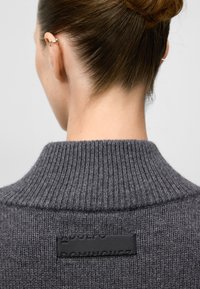 Close-up van de nek en rug van een persoon die een grijze geribbelde trui draagt met een zwart leren label dat op de bovenrug is genaaid.