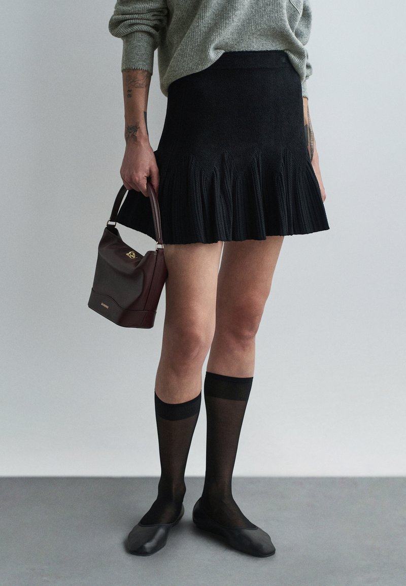 Schwarzer, plissierter Minirock mit strukturierter Oberfläche, kombiniert mit durchsichtigen schwarzen Socken und schwarzen Ballerinas, hält eine bordeauxfarbene Handtasche.
