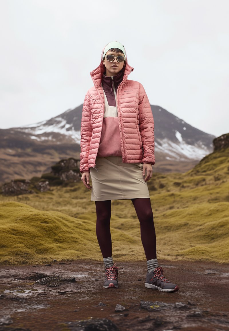 Giacca a vento rosa con cappuccio, indossata sopra un top color bordeaux. Gonna beige, collant bordeaux e scarpe da trekking grigie con dettagli arancioni. Sfondo montano.