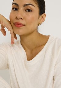 Top de manga larga texturizado en color beige claro con un escote amplio. Modelo con un moño estilizado, pendientes de aro dorados descansando su barbilla sobre la mano.