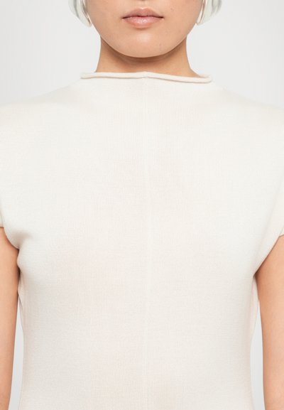 Persoon in een witte mouwloze gebreide top met hoge kraag en over-ear witte koptelefoon, getoond van kin tot schouders.