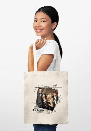 Femme souriante aux cheveux noirs tenant un sac tote beige avec une photo du casting de Gossip Girl et le slogan « La popularité a un prix. »