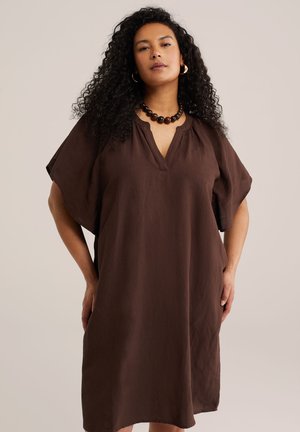 Žena s kučeravými tmavými vlasmi, oblečená v voľných hnedých šatách, s čiernym náhrdelníkom z korálikov a zlatými kruhovými náušnicami, stojaca s rukami po bokoch.