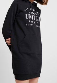 Robe sweatshirt noire à capuche avec poche avant, ornée d'un texte imprimé blanc : "LOS ANGELES UNITED COSMIC AERONAUTICS." Matière douce, coupe décontractée.