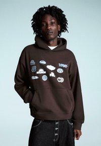 Brązowa bluza z kapturem ozdobiona wieloma niebiesko-białymi logo, posiadająca kieszeń kangurkę i regulowany kaptur na sznurek, zestawiona z ciemnymi dżinsowymi spodniami.