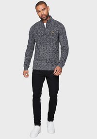 Grå og sort strikket sweater med høj krave og ribbede manchetter, der har et lille gult logo accent på brystet. Båret med sorte slim-fit jeans.