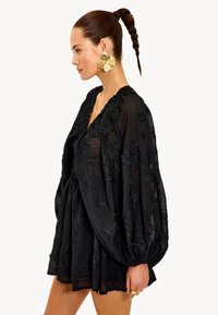 Vestido negro con patrones florales texturizados, mangas voluminosas y un escote con volantes. Tejido ligero y transparente con un corte holgado.